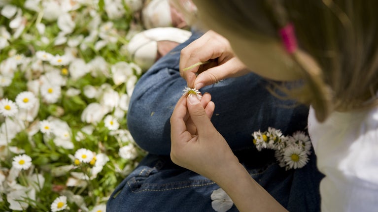 Close-up of a child sitting in a grassy meadow filled with white daisies, carefully threading flowers together to make a daisy chain on a sunny day.
